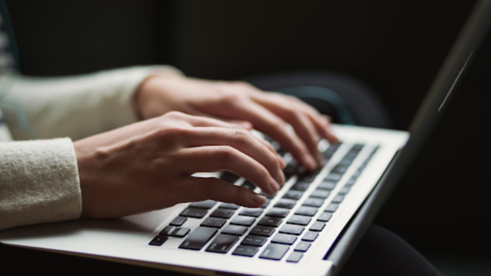 Hands typing on a laptop keyboard in a dimly lit setting.